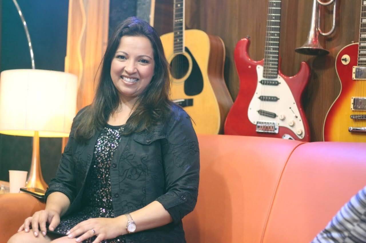 Adriana Junior sentada em um sofá laranja, sorrindo, diante de uma parede coberta de violões e guitarras.
