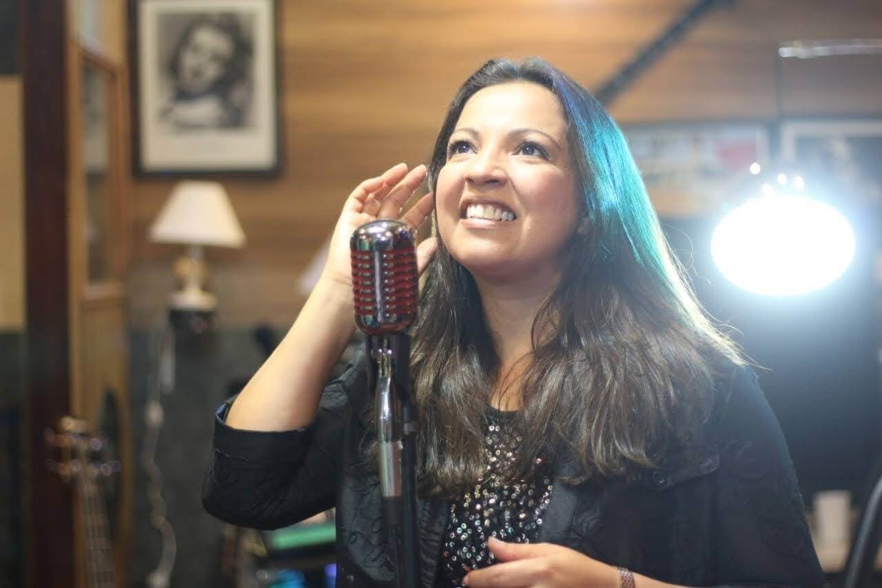 Adriana Junior diante de um microfone vintage em estúdio, sorrindo e olhando para cima.