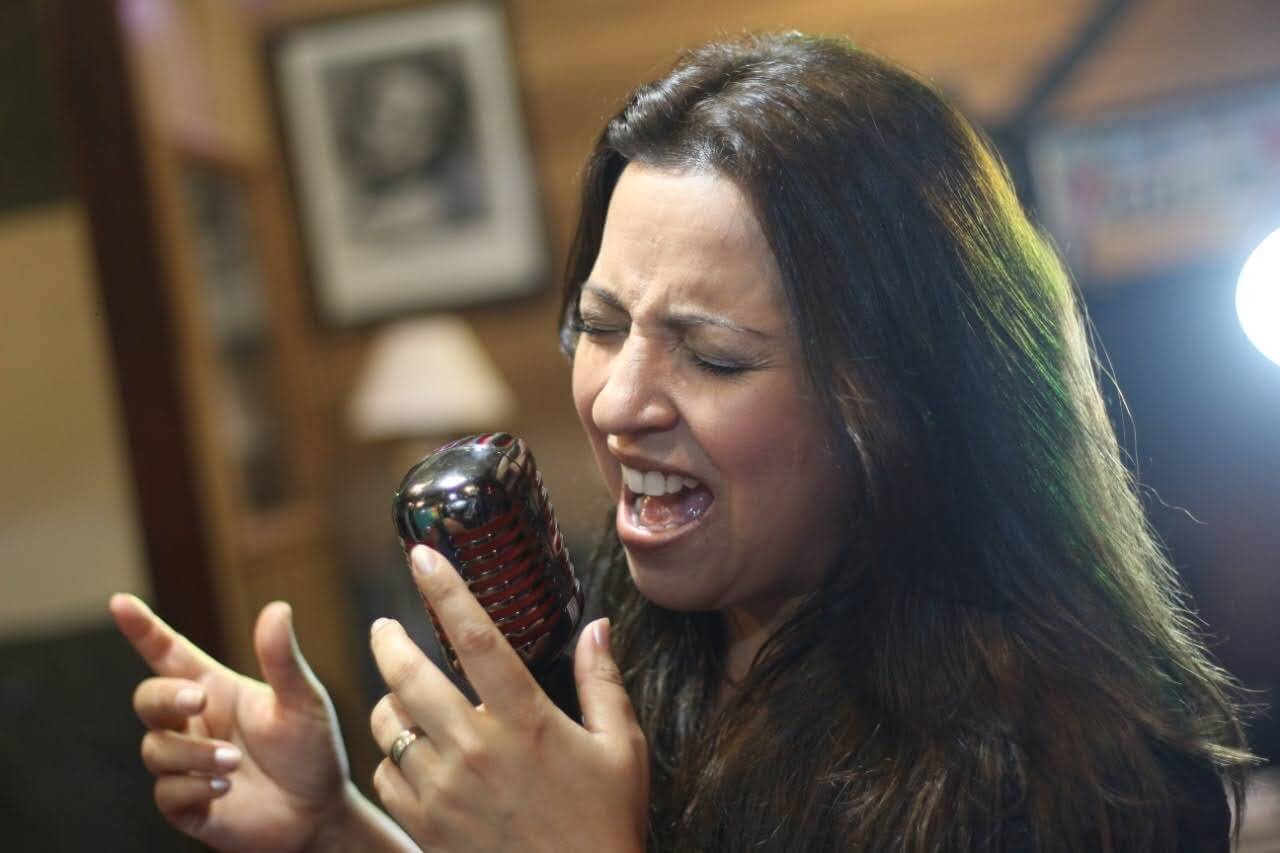 Close de Adriana Junior cantando com intensidade em um microfone vintage.