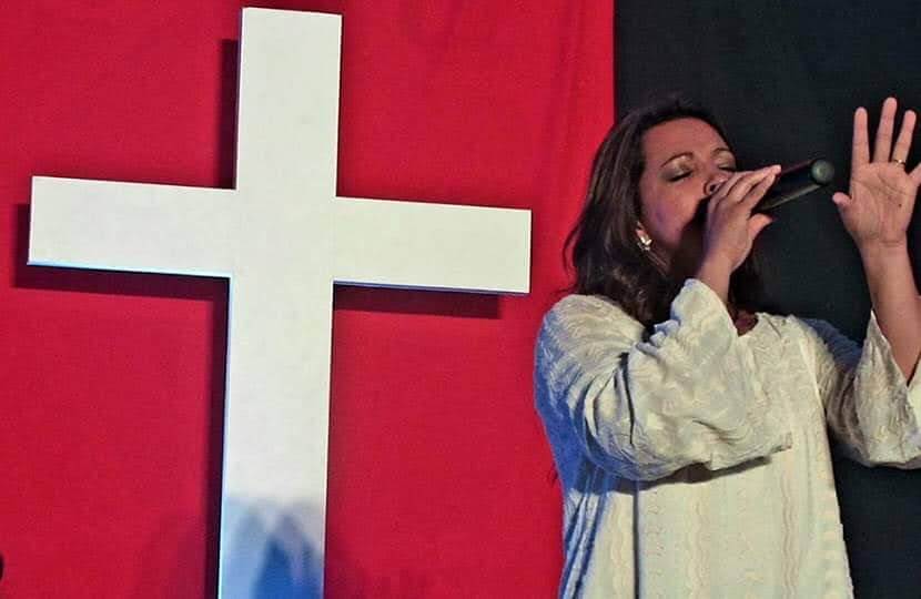 Adriana Junior cantando de olhos fechados ao lado de uma grande cruz branca, sobre fundo vermelho.