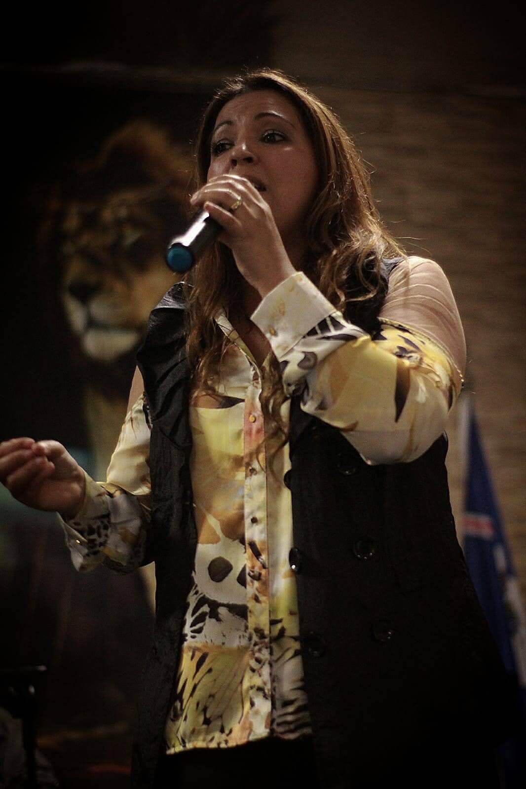 Adriana Junior cantando ao microfone durante uma apresentação ao vivo, sob luz quente de palco.